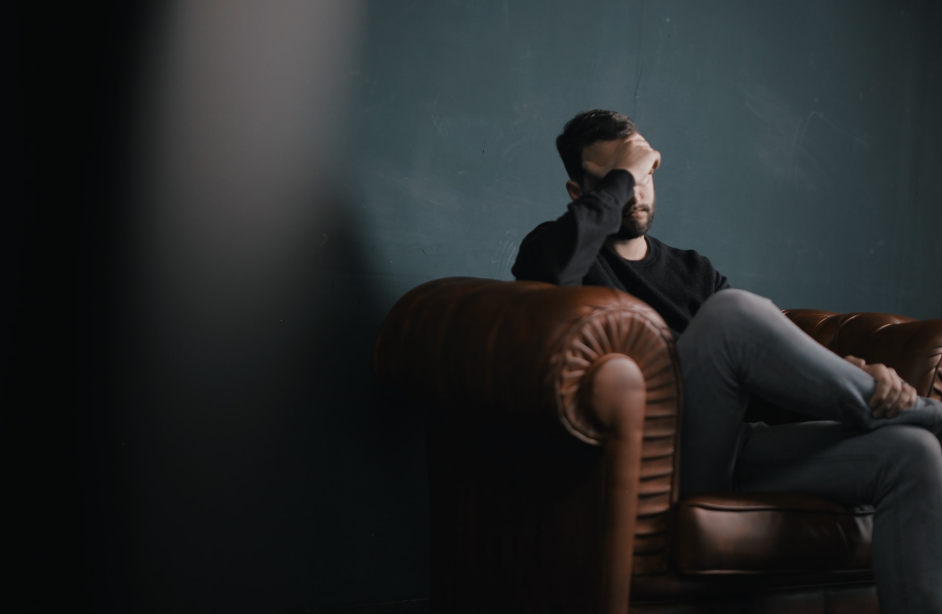 Man sits on brown leather sofa with hand rested on forehead