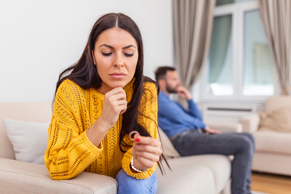 woman with long brown straight hair in yellow sweater holding wedding ring sitting on couch. man in foreground.