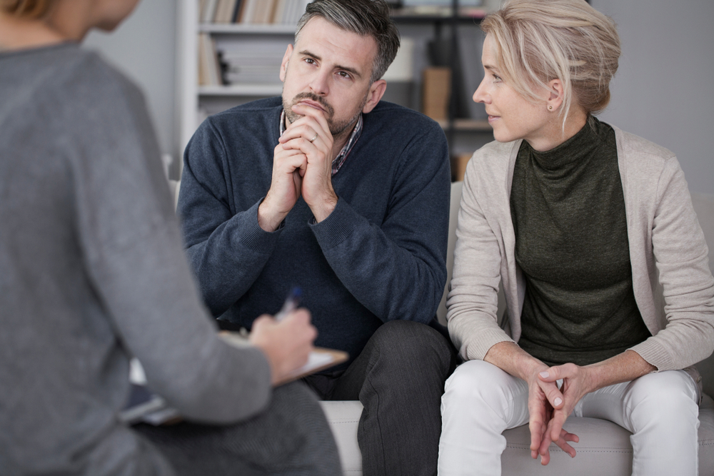 man with navy sweater sitting next to woman in green turtleneck on couch. they are speaking to a couples counselor.