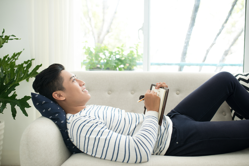young man at home writing writing down thoughts in journal on notebook, sitting on couch